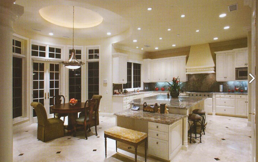 Spacious white kitchen with island, dining area, and large windows. Beige tile floor, neutral walls.