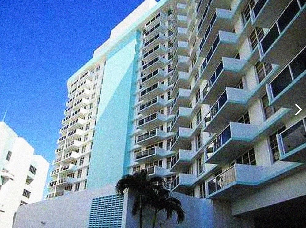 Blue and white high-rise building with balconies, likely a hotel or condo, against a clear sky.