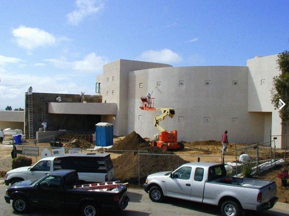 Construction site with white building, trucks, and equipment under blue sky.