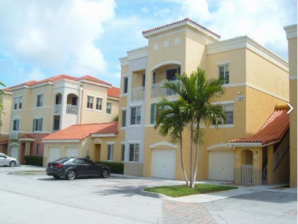 Buildings with beige stucco walls, red tile roofs, and attached garages, with a palm tree in front.