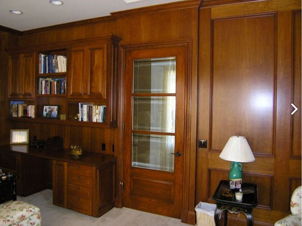Wooden paneled office with a desk, bookshelves, and a glass-paned door. A lamp sits on a small table.