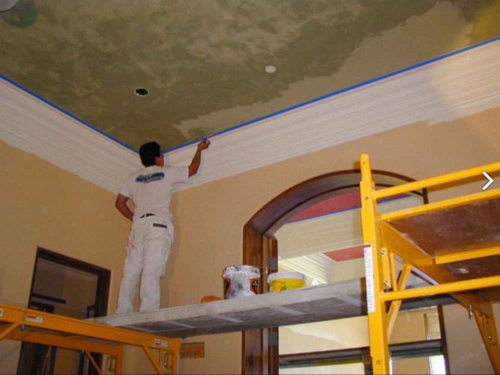 A person painting a white ceiling molding. They stand on a scaffold, using a brush, inside a room.