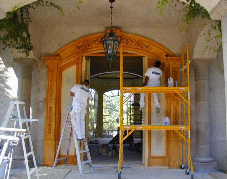 Two painters on ladders applying finish to a large ornate wooden doorway and surrounding structure.
