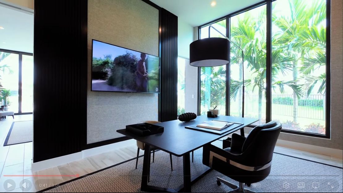 Office with desk, TV, and large windows overlooking palm trees. Black furniture, light colored walls and rug.