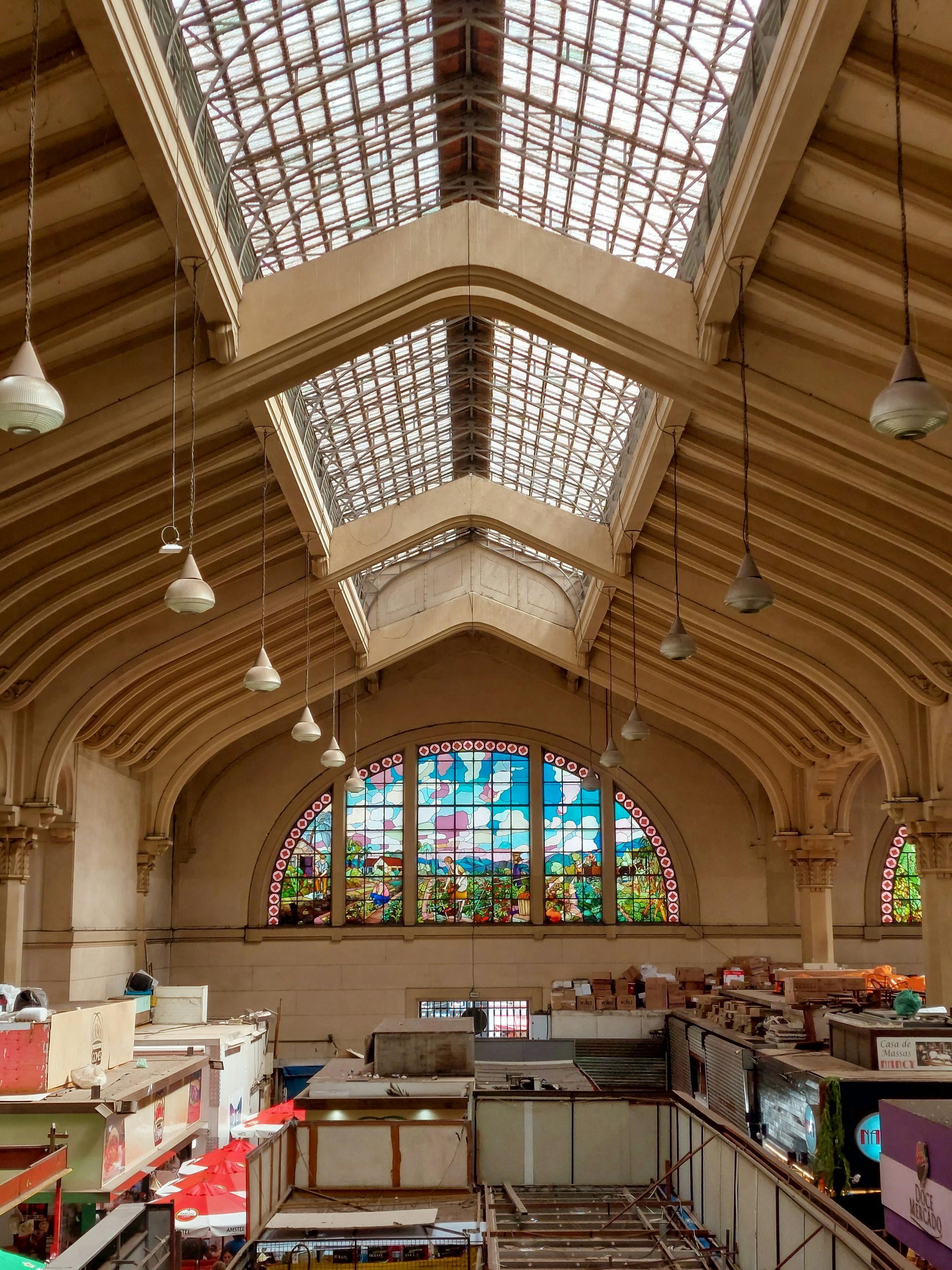Inside of a large hall with glass ceiling and stained-glass window. Brown wooden beams, white lights, and shop counters are visible.