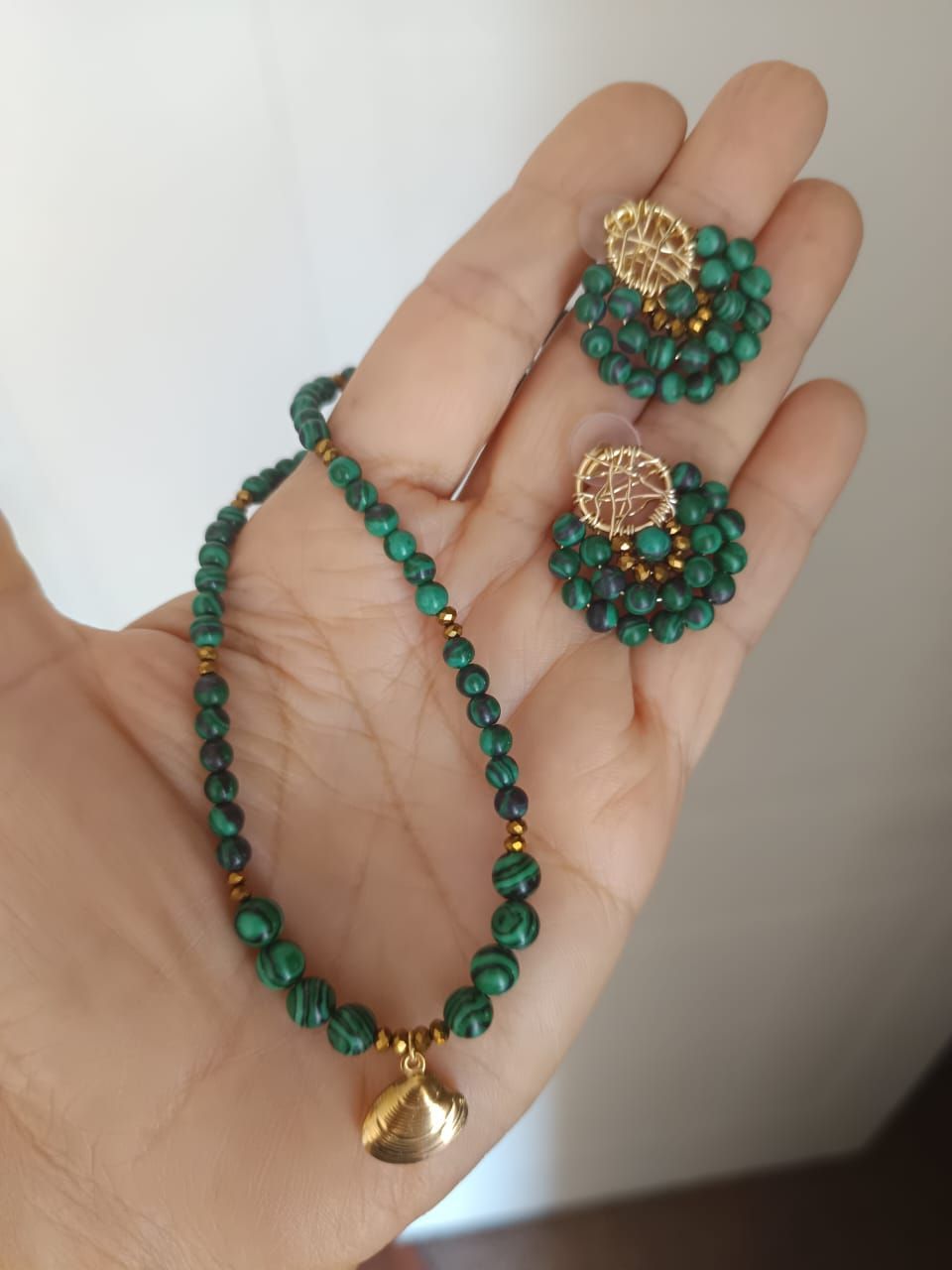 Una mano sostiene un collar de cuentas de malaquita con un dije de concha de oro y pendientes a juego de cuentas de oro y malaquita.