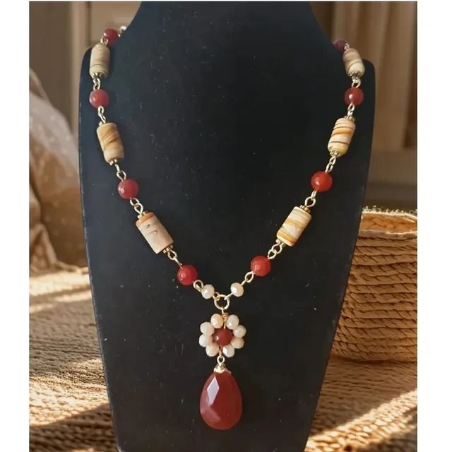 Un collar de cuentas con piedras cilíndricas estampadas, cuentas esféricas rojas y un dije central en forma de flor con un colgante en forma de lágrima.