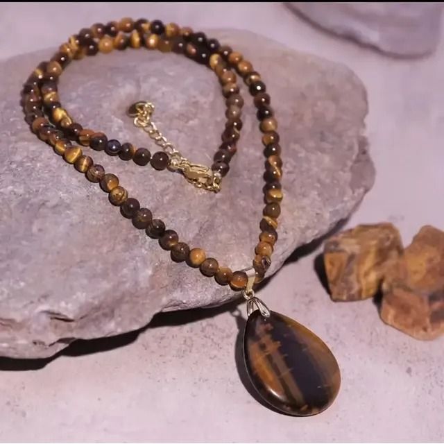 Un collar con cuentas de ojo de tigre y un gran colgante pulido en forma de lágrima, que descansa sobre una superficie de piedra gris texturizada.