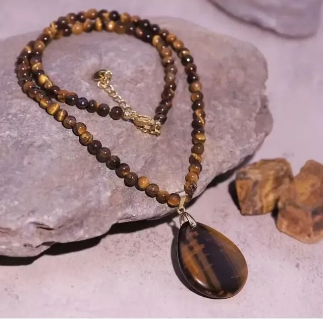 Un collar hecho de cuentas de ojo de tigre marrones y rayadas con un gran colgante en forma de lágrima, engastado sobre una superficie de piedra texturizada.