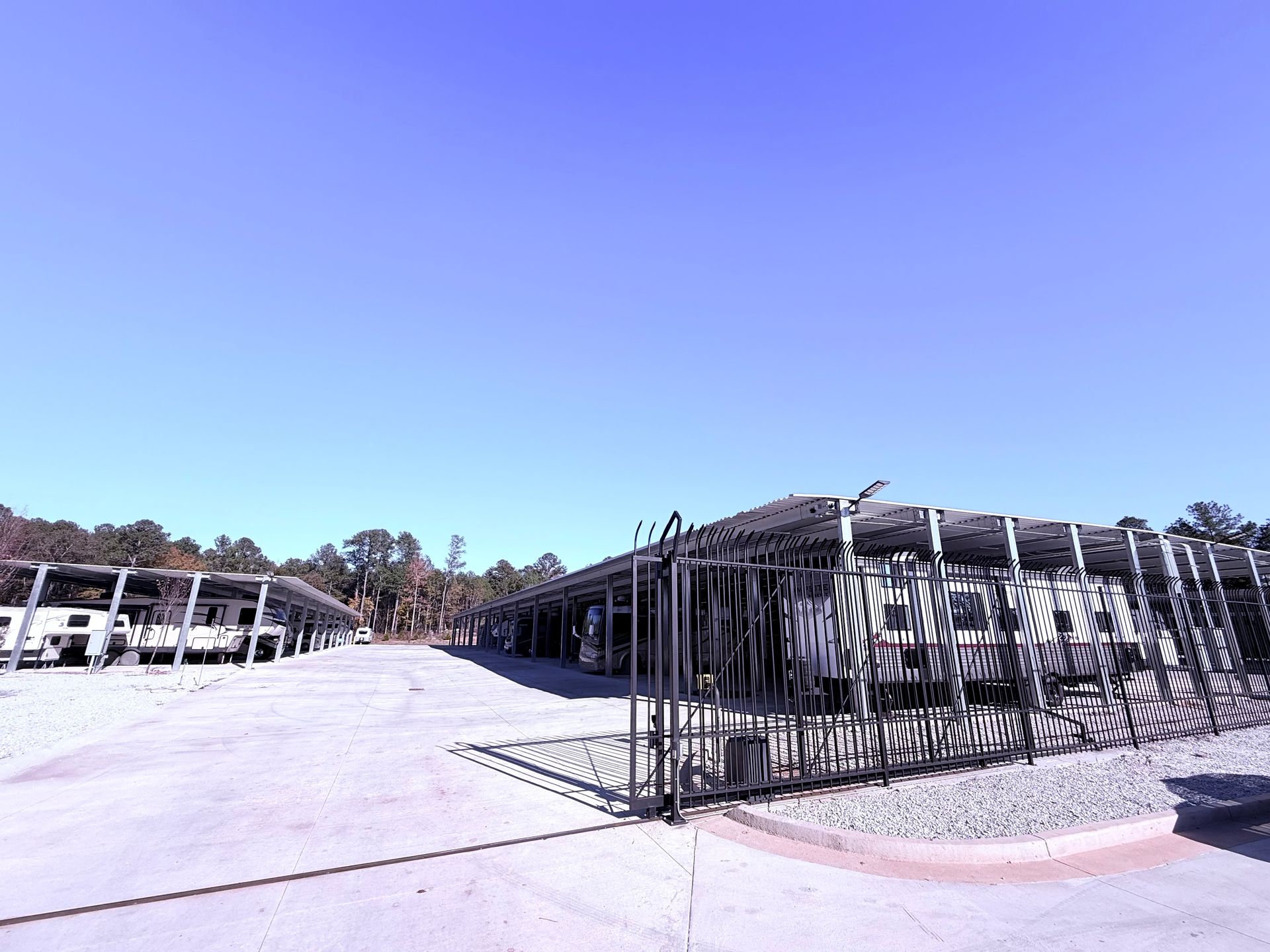 Gray parking lot with covered carports under a blue sky; a black gate is in the foreground.