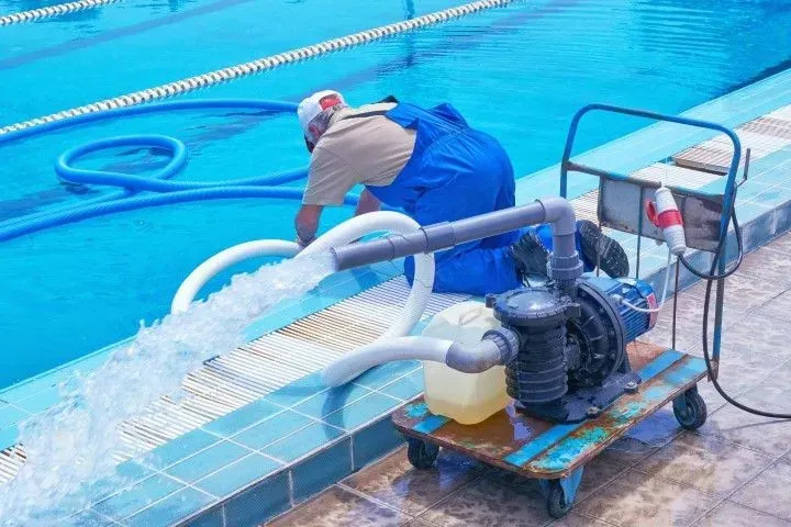 Worker using a pump to drain bright blue pool water onto a wet deck