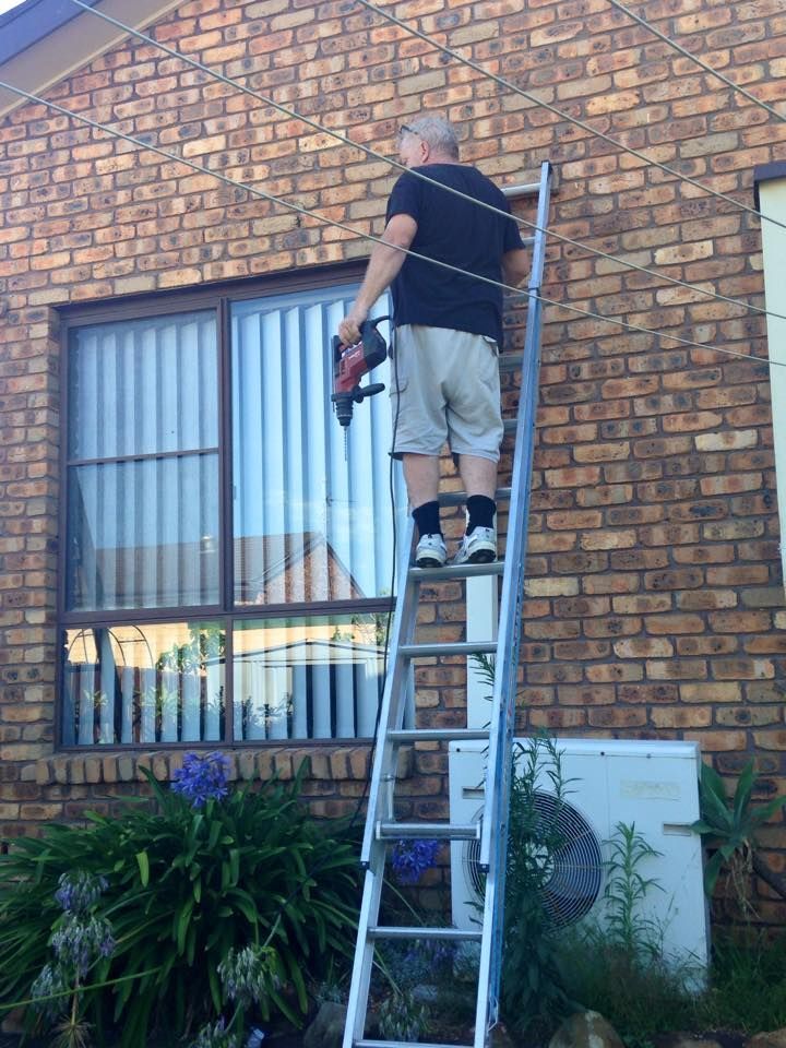 A Man is Standing on a Ladder in Front of a Brick Building — Design 2000 Blinds & Awnings In San Remo, NSW