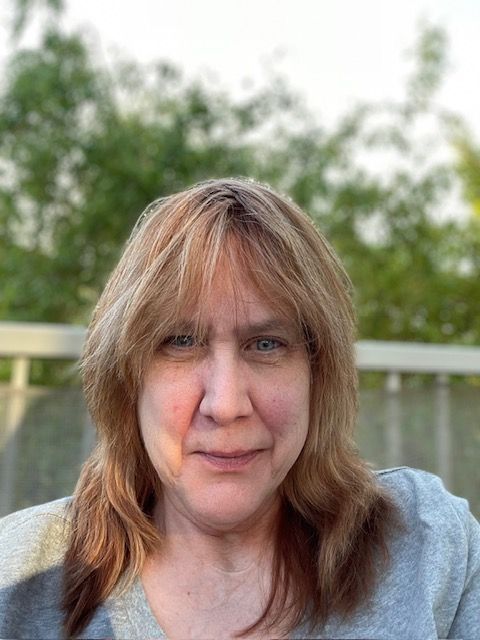 A person with shoulder-length, light brown hair and bangs looking forward, set against a blurred outdoor background.