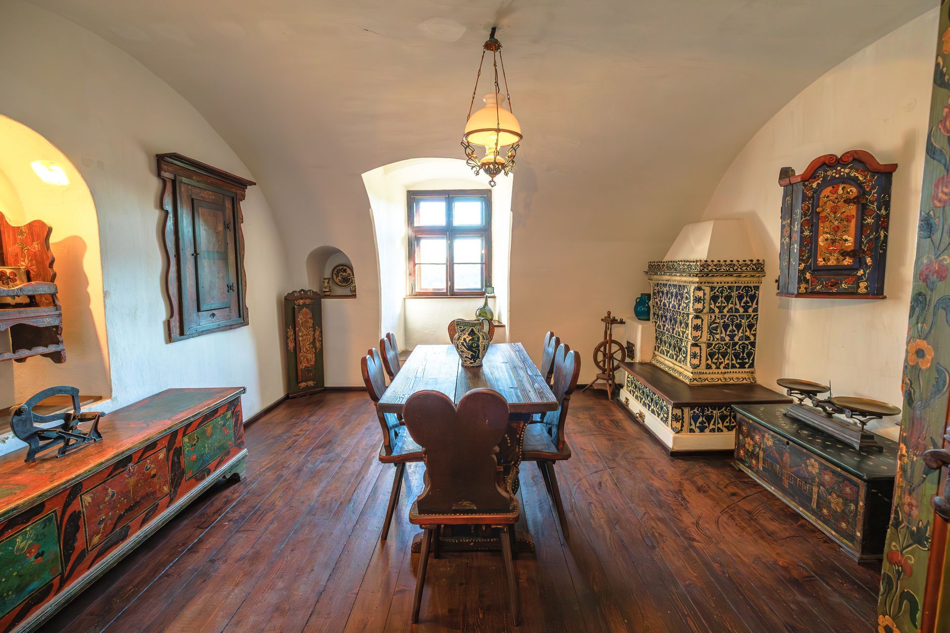 Dining room with wooden table, chairs, decorative chests, and ornate fireplace, with a window and a hanging light.