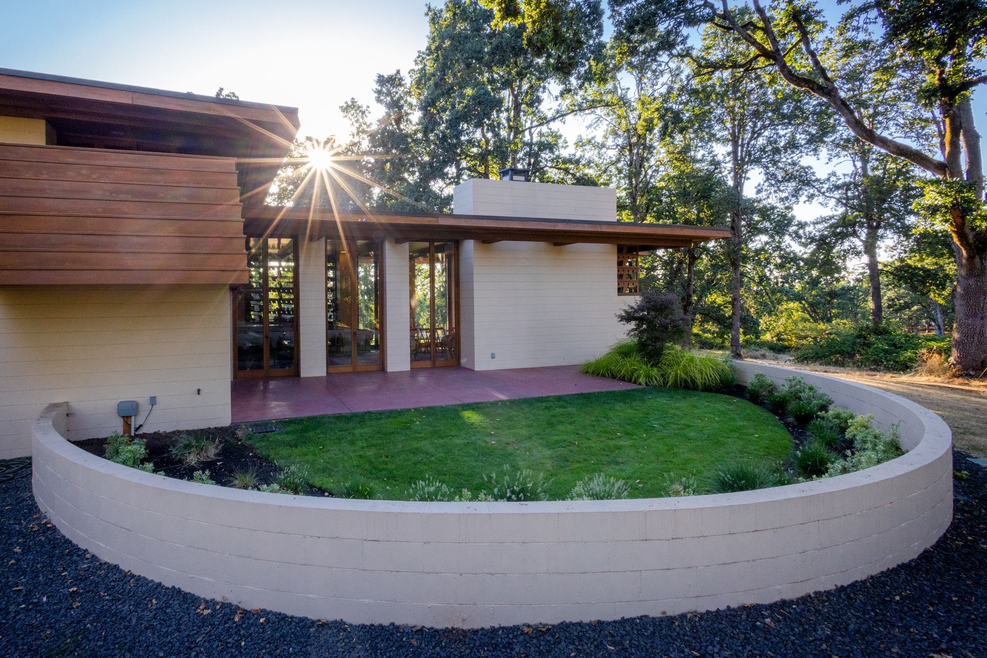 Modern home with a curved wall around a small lawn; sunlight shines through the trees.