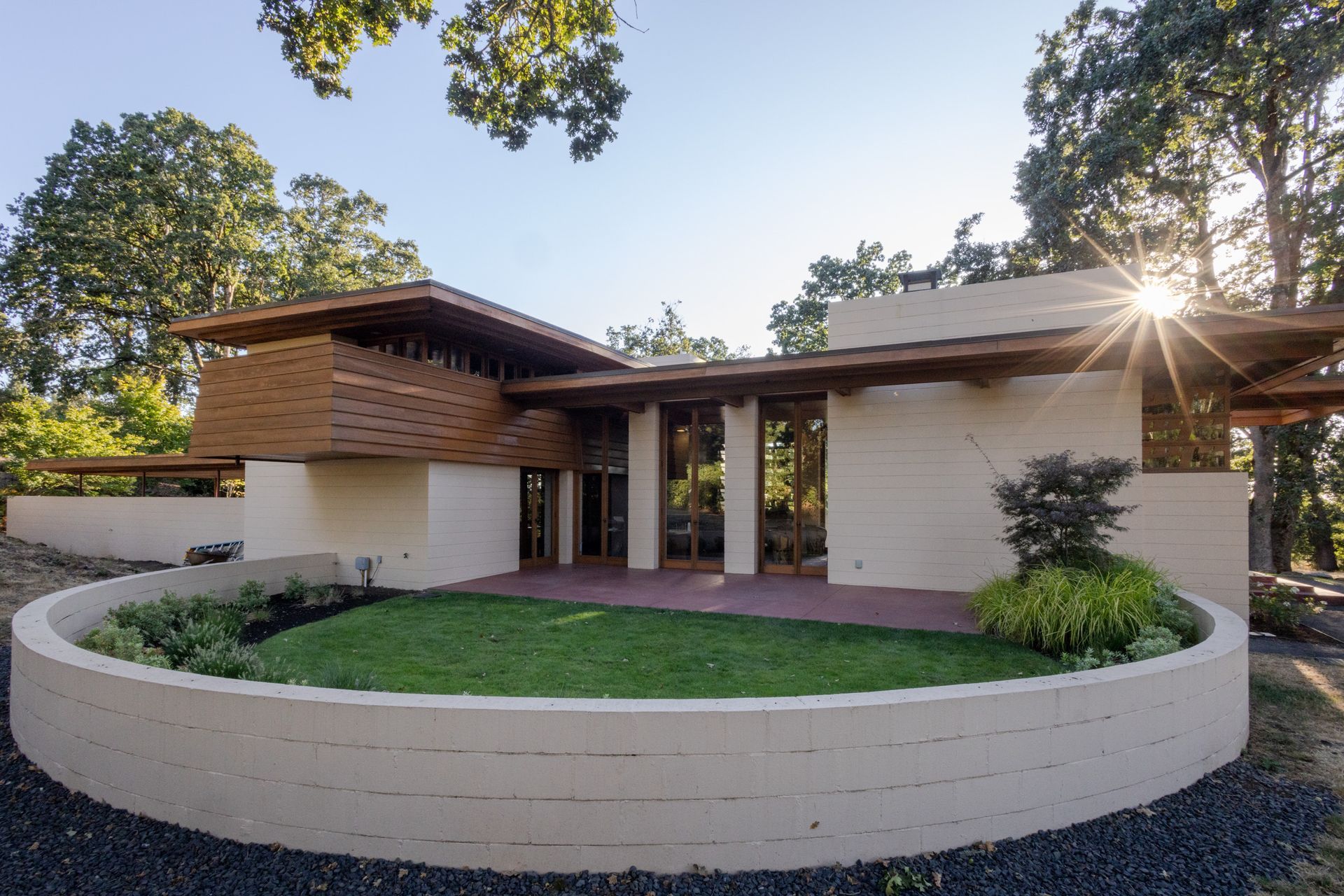 Modern, low-slung house with light-colored walls, wood trim, and a circular lawn area. Sunburst in the background.