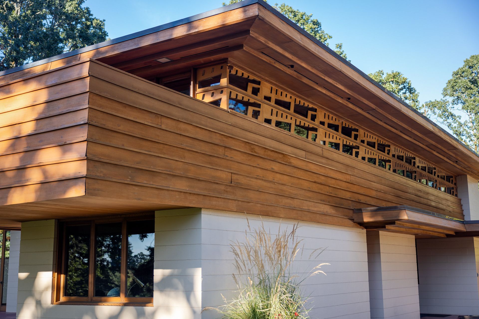 Two-story wooden house with open-air balcony, cream-colored walls, and brown accents. Sunlight casts shadows.