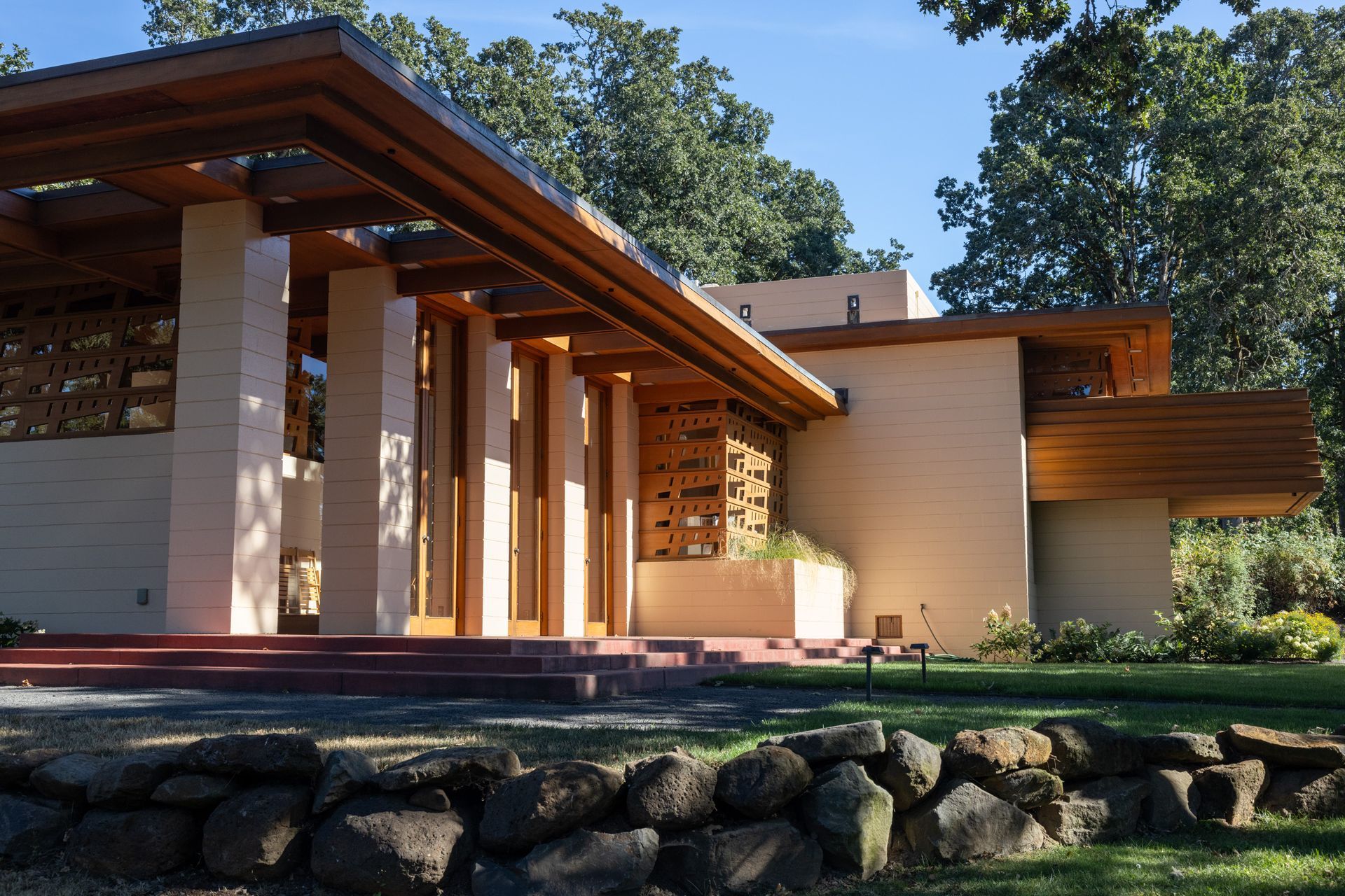 Beige brick house with wooden beams and columns. Landscaping includes rocks and grass.