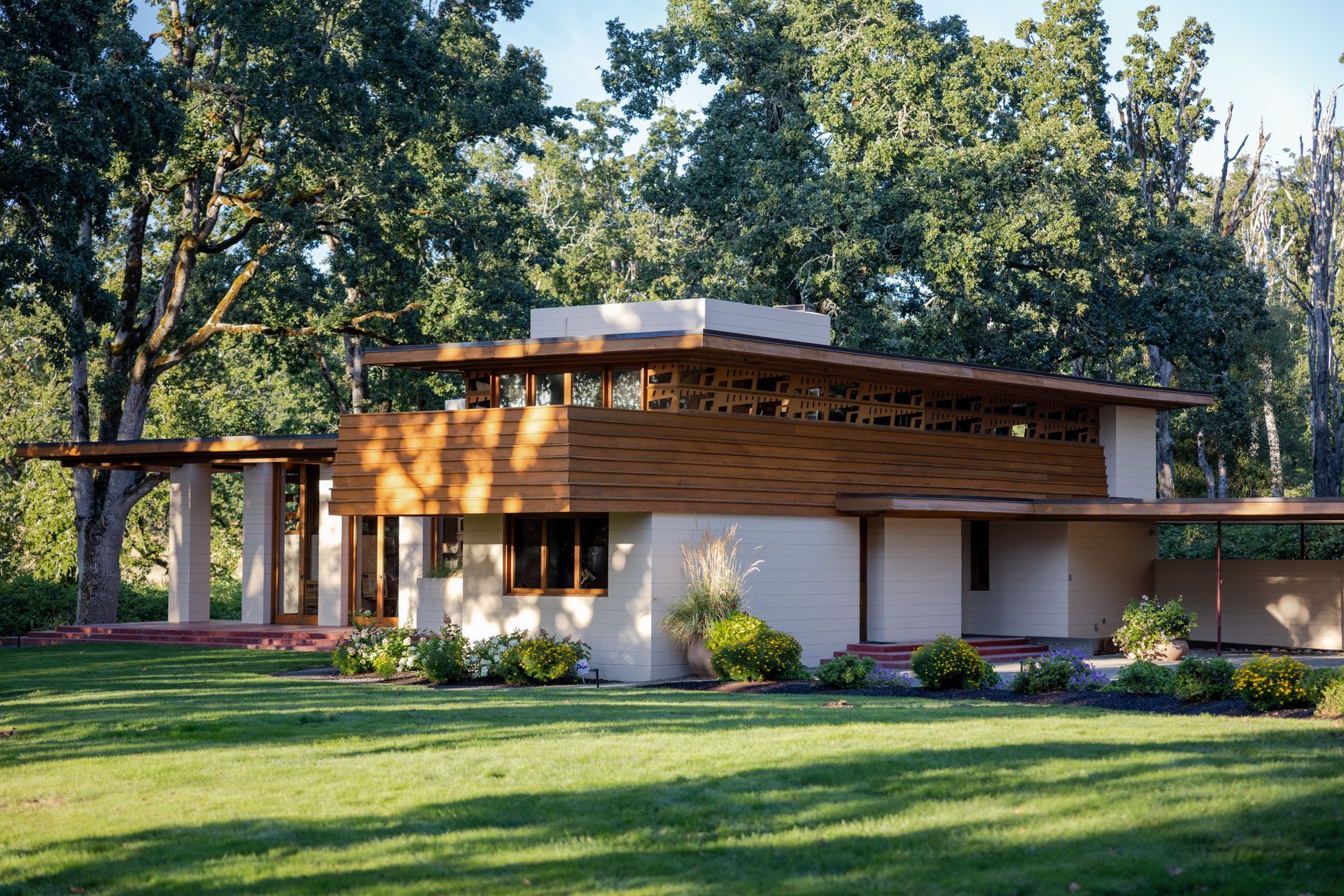 Modern, single-story house with flat roof, horizontal wood accents, and light-colored walls, surrounded by trees and grass.