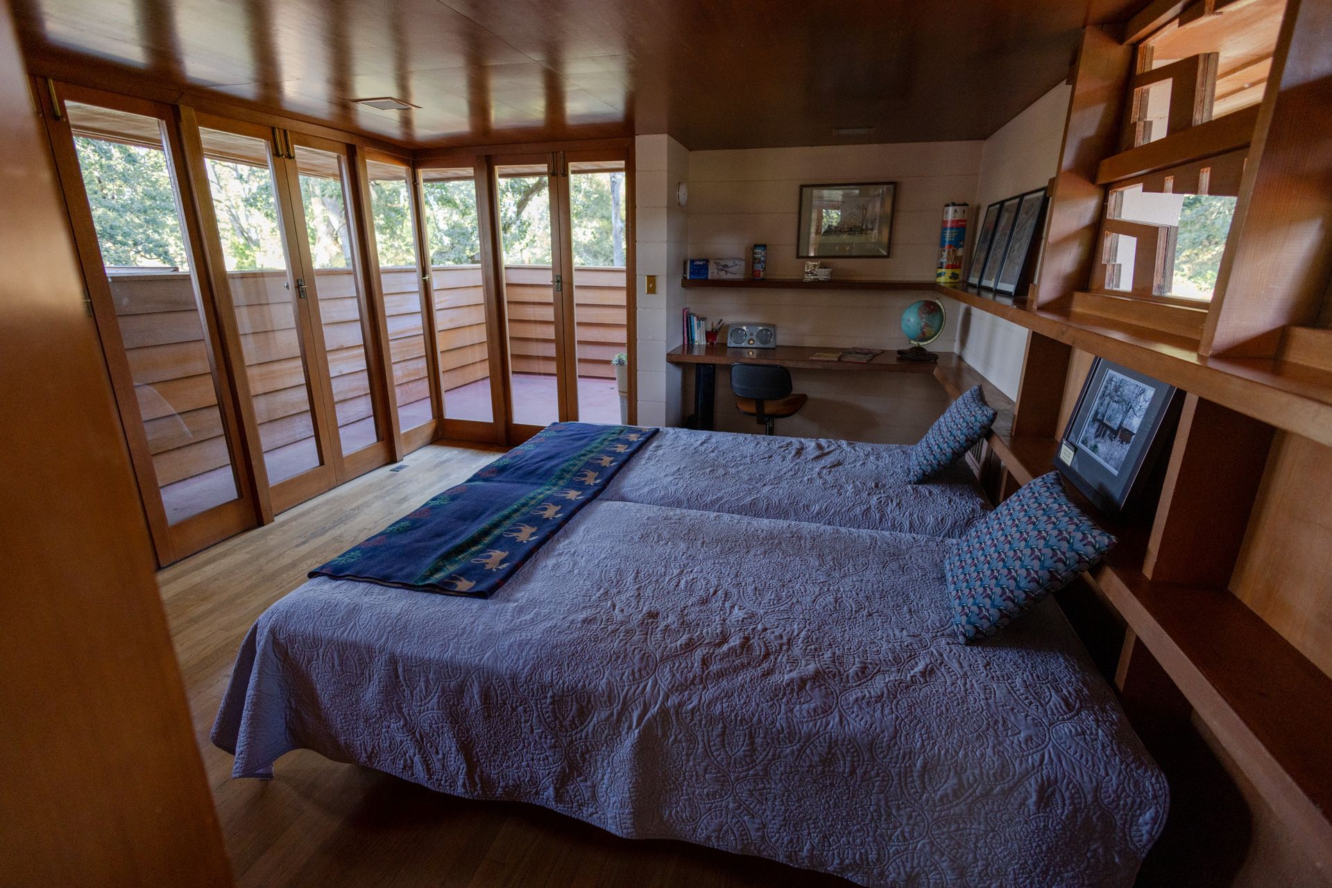 Bedroom with two beds, wooden built-in shelving, desk, and large windows.
