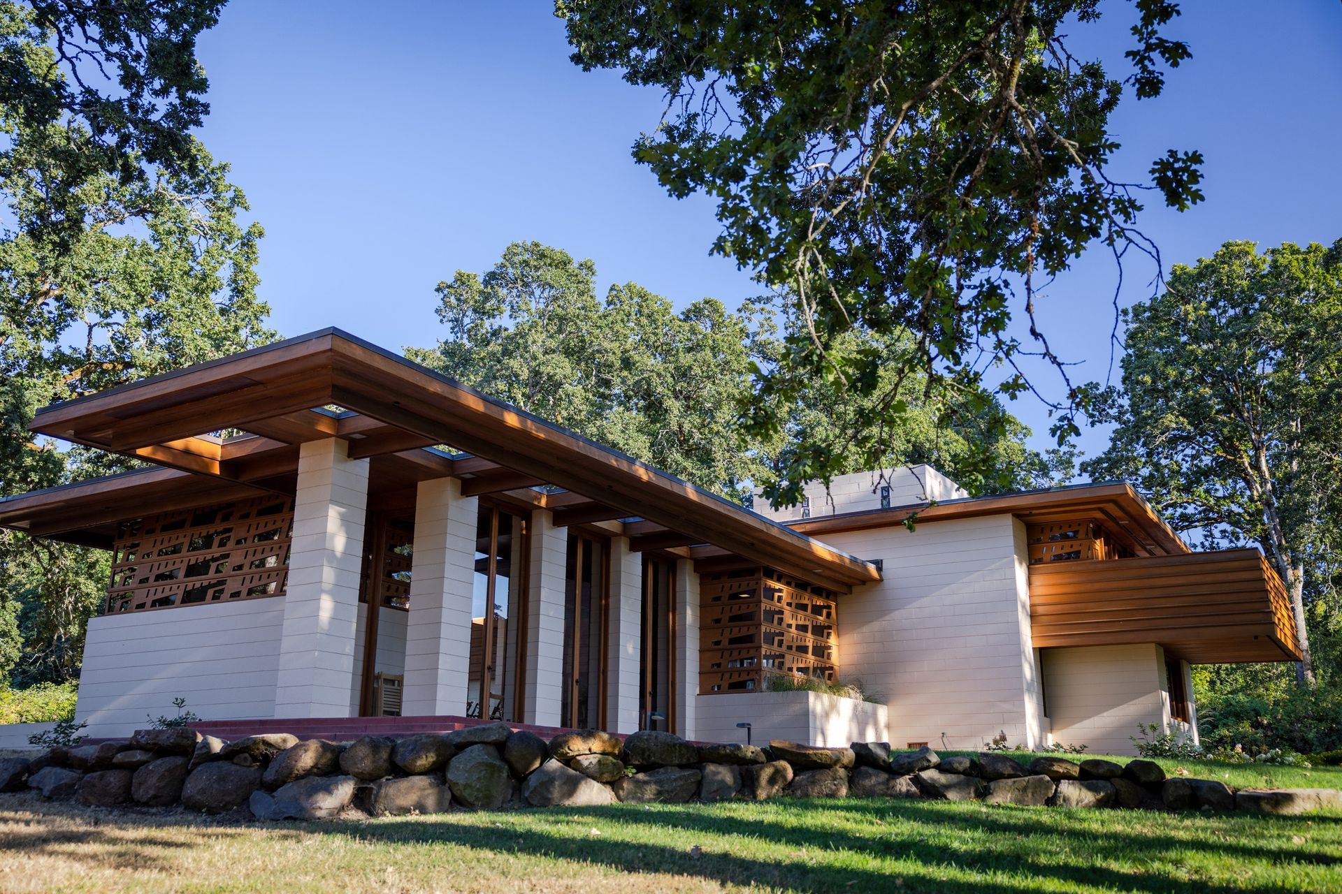 A low, white brick house with a wooden roof and beams, set in a grassy yard under a blue sky.