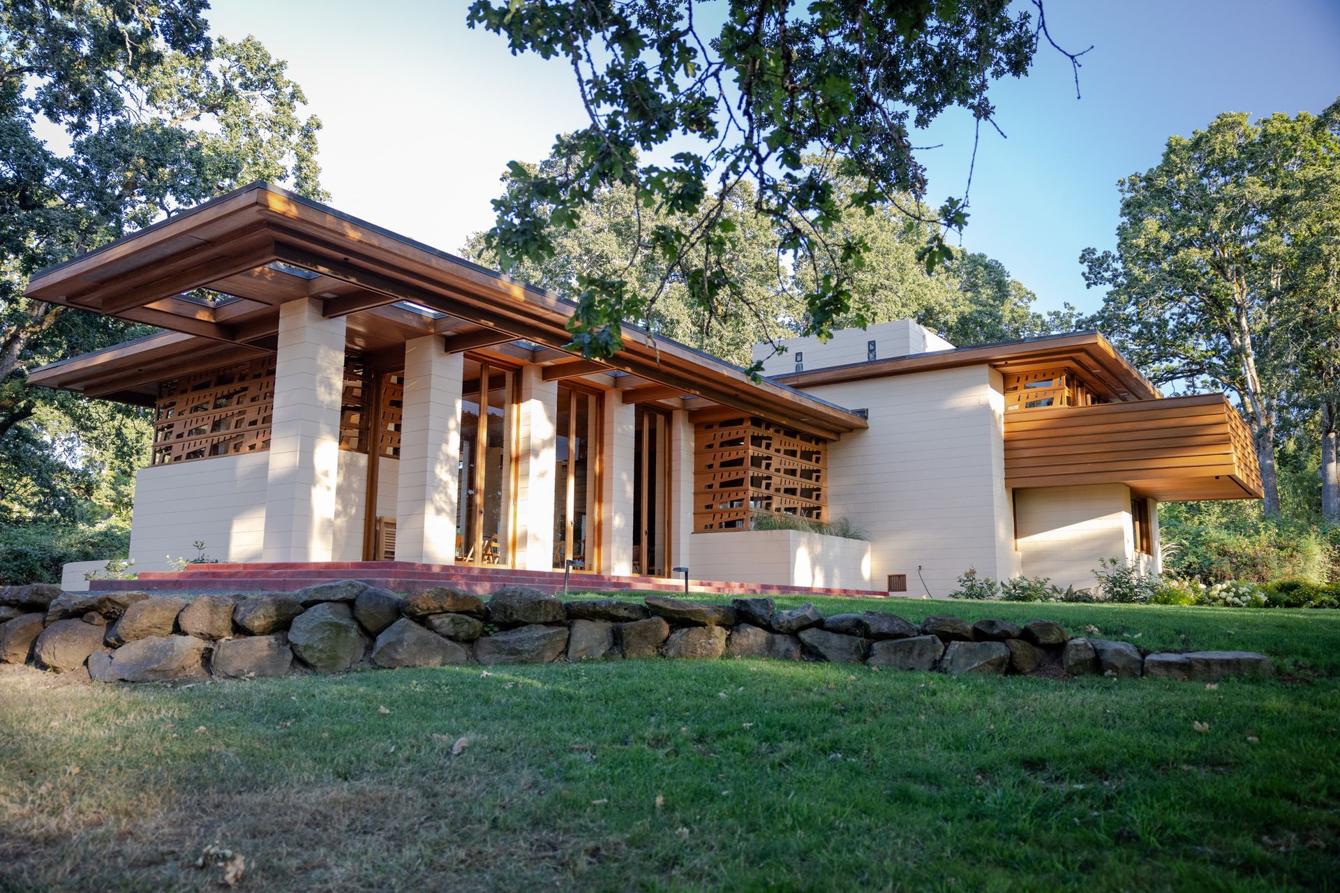 Frank Lloyd Wright-designed house with light tan walls, wood accents, and overhanging roof. Set on a grassy hill with stone wall.