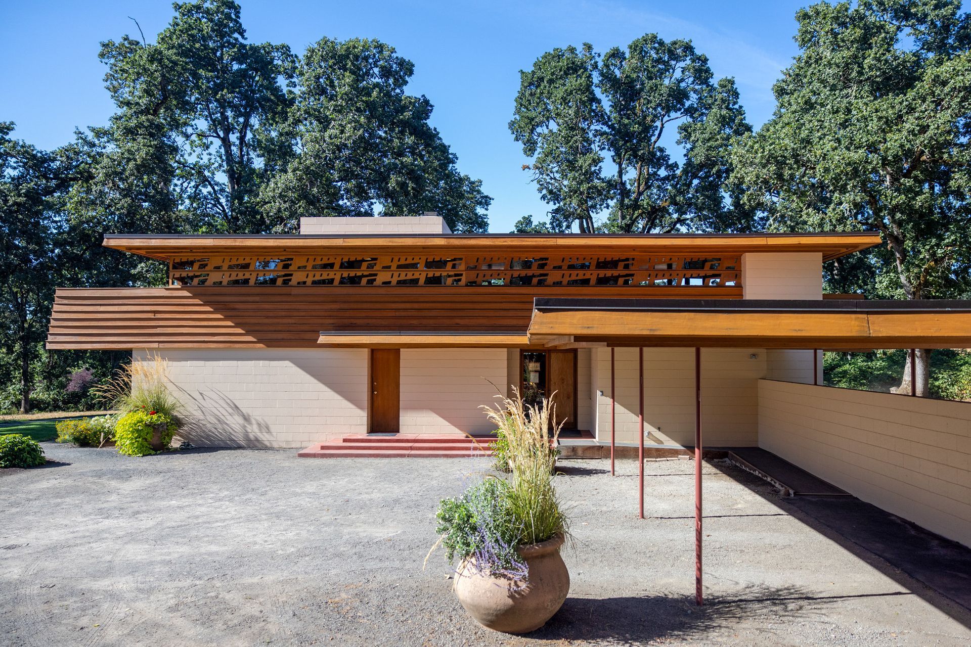 Modern, single-story home with a flat roof, horizontal lines, and wooden accents. Gravel driveway and trees in the background.