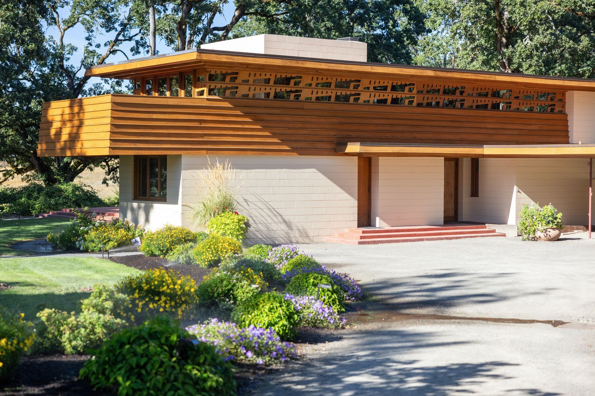 Low-slung, light-colored modern house with wooden trim, surrounded by landscaping and a driveway.