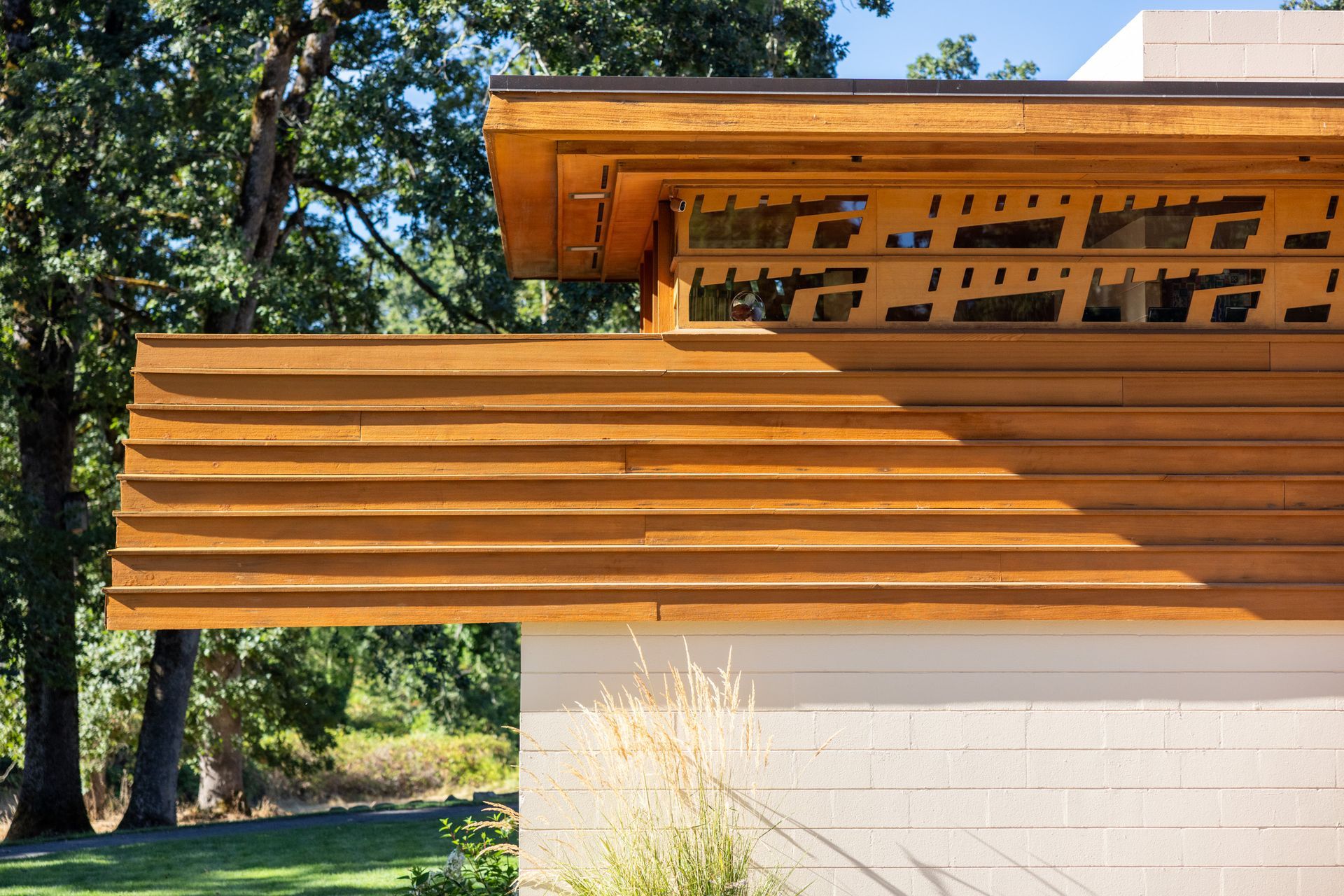 Wooden roof overhang with patterned details, against a white stucco wall and trees.