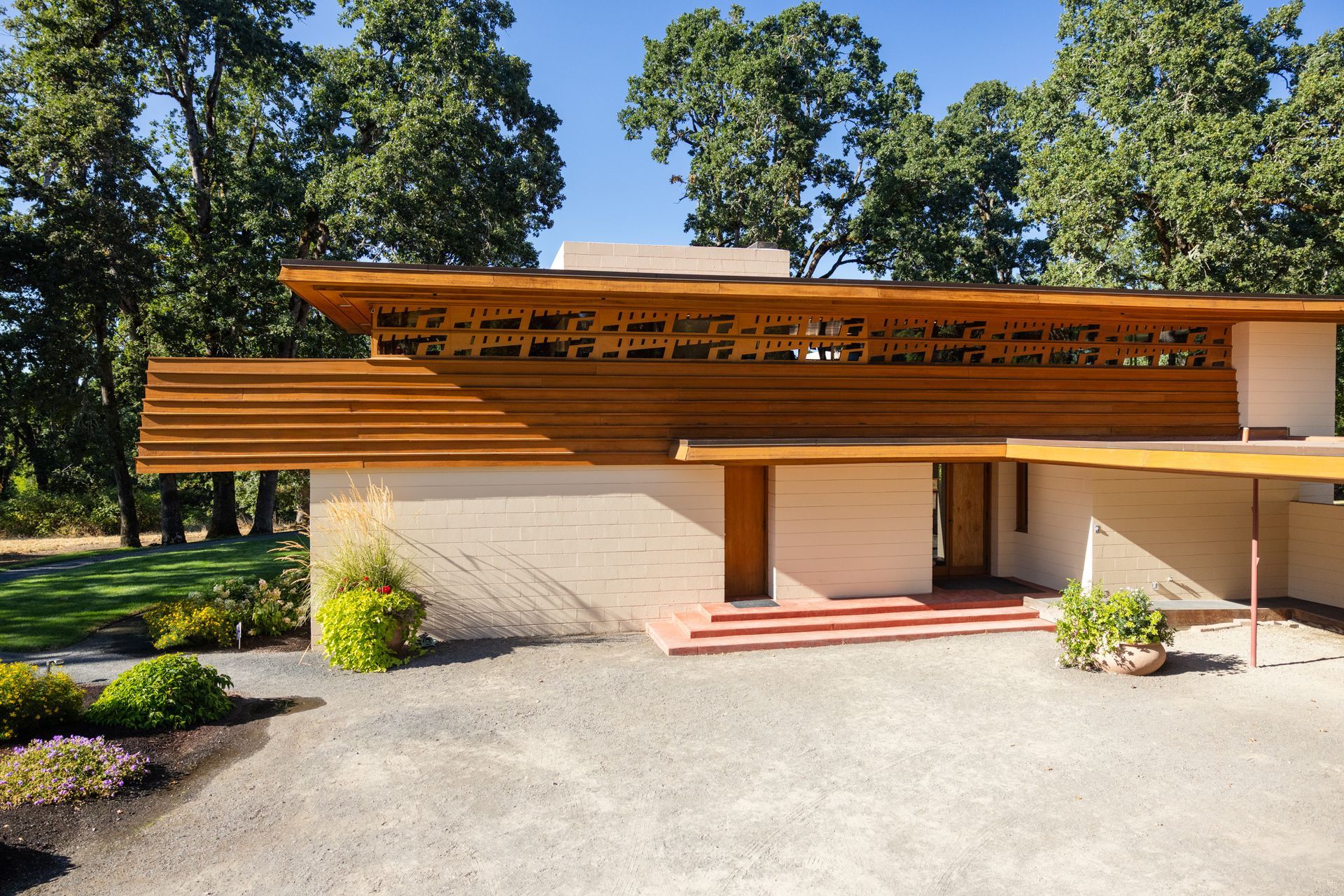 Low-slung, light-colored house with wooden accents and a carport, trees in background, sunny day.