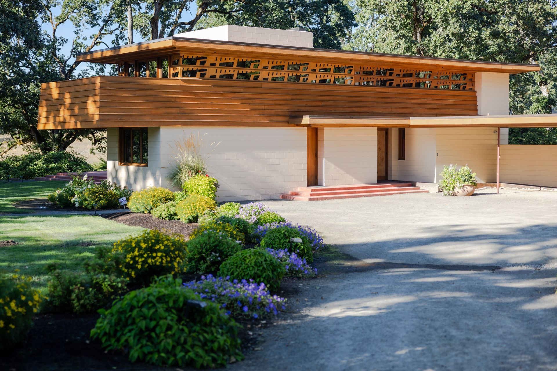 Low-slung, cream-colored home with wooden accents. Landscaping includes green bushes and purple flowers.