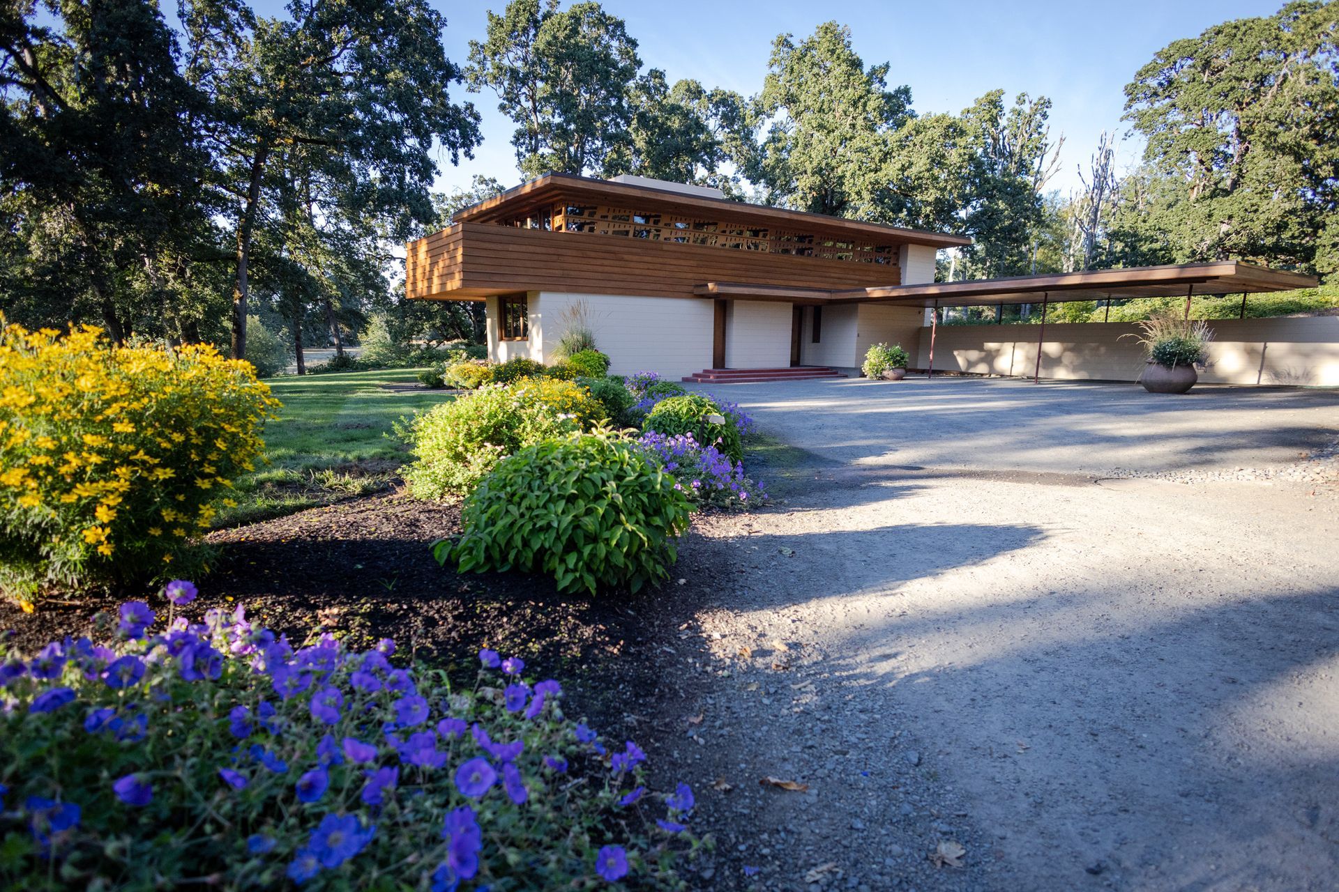 Low-angle view of a beige and brown house with landscaped garden and gravel driveway under trees.