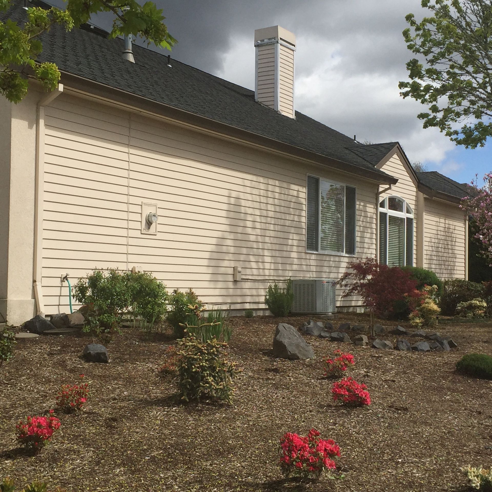 Tan-sided house with dark roof, chimney, and landscaping. Cloudy sky overhead.