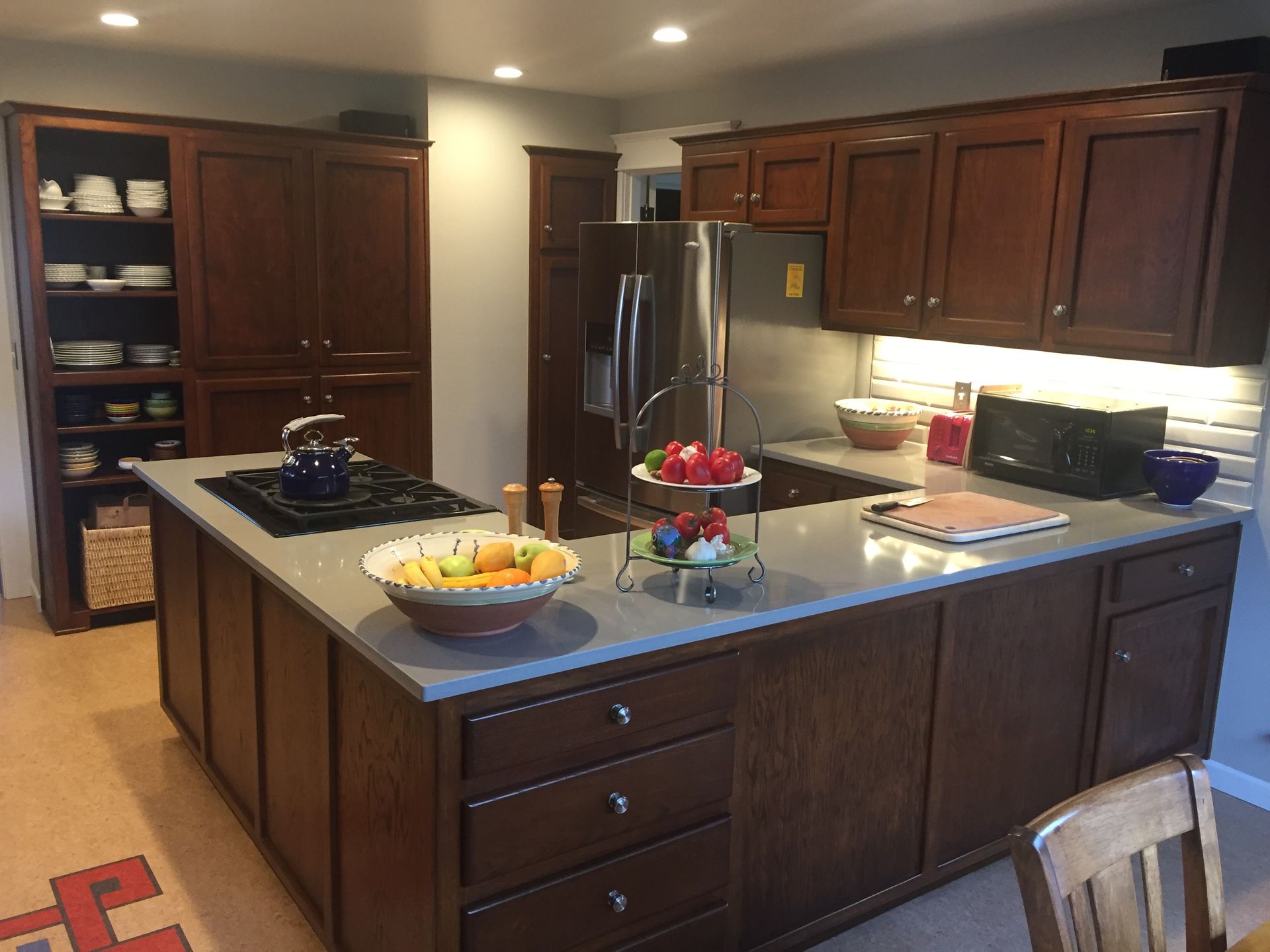Kitchen with wooden cabinets, island with a stovetop, and a refrigerator. Fruits on countertop.