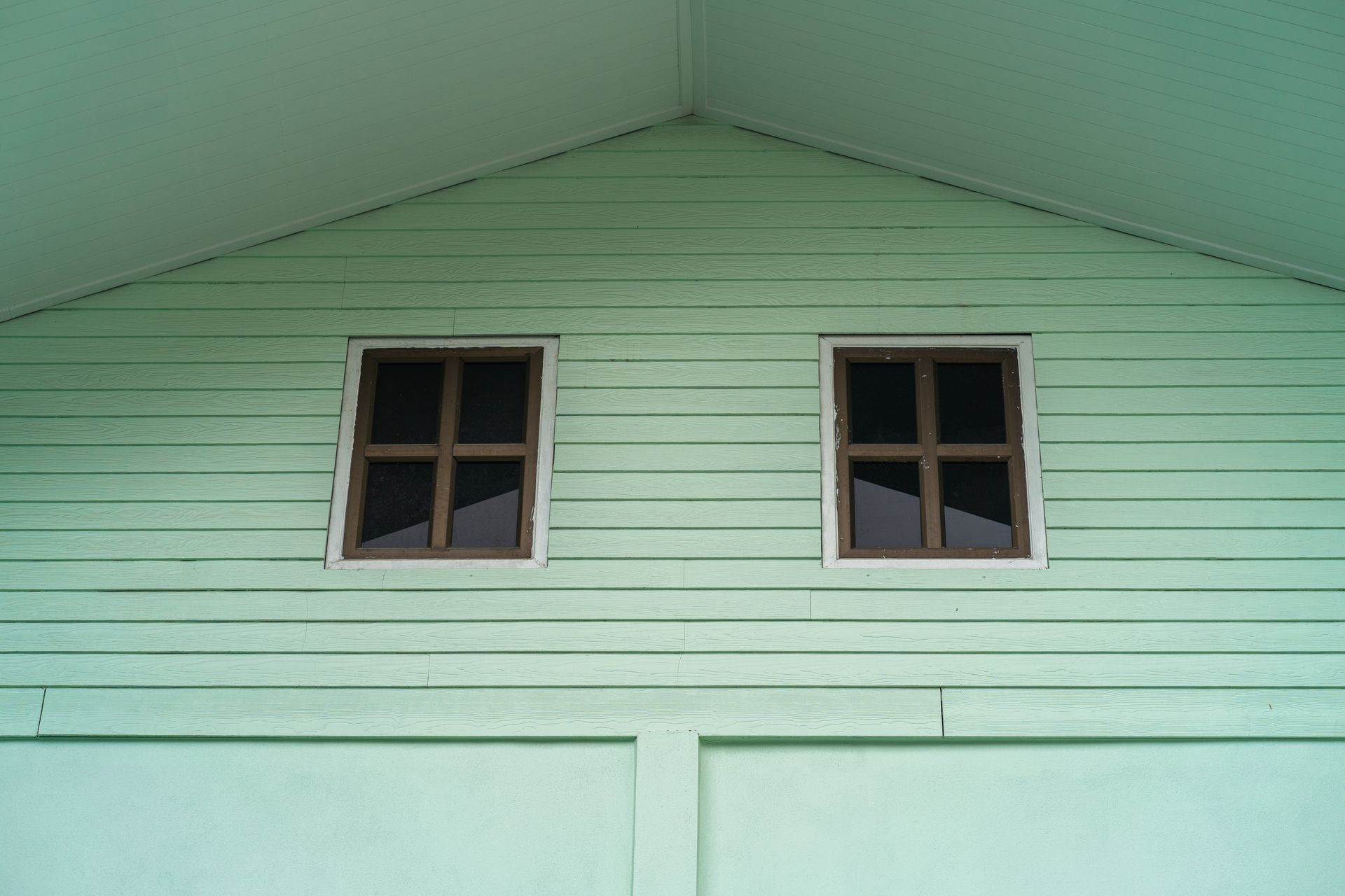 Green building with two windows under a roof, brown window frames, and horizontal wooden siding.