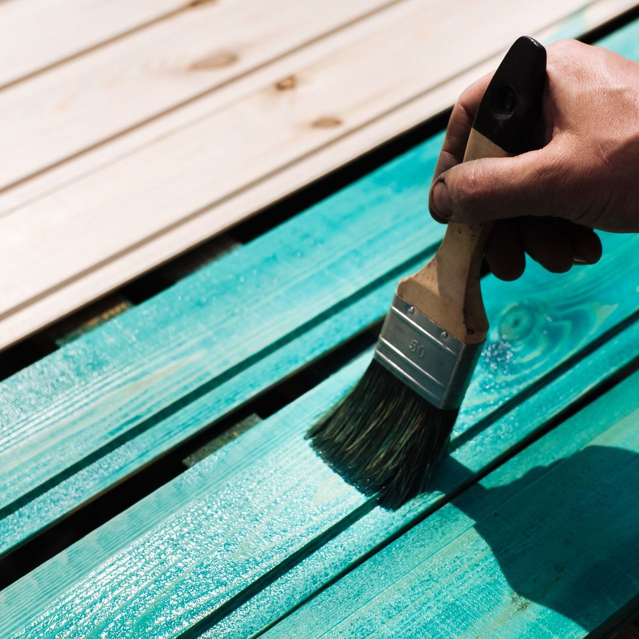Hand painting teal paint on wooden planks with a paintbrush outdoors.