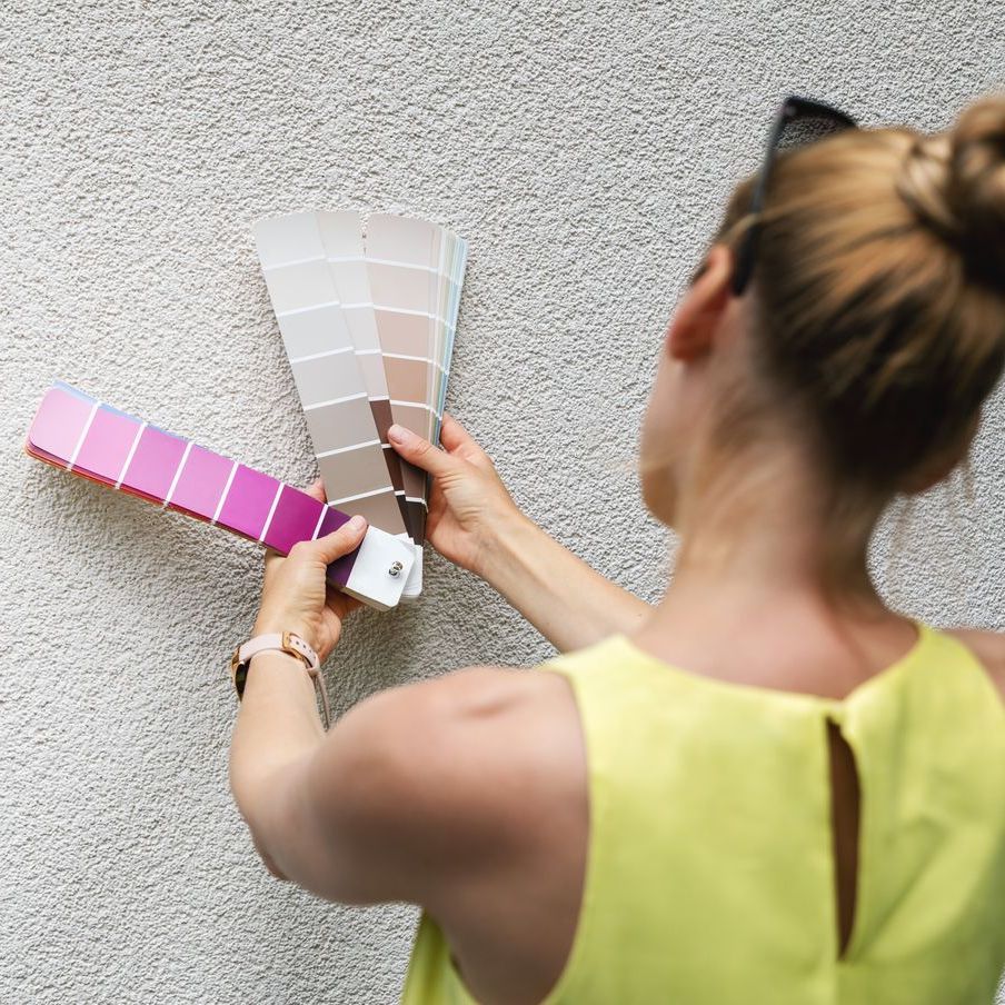 A woman holds up paint swatches against a textured wall. She wears a yellow top and has her hair in a bun.