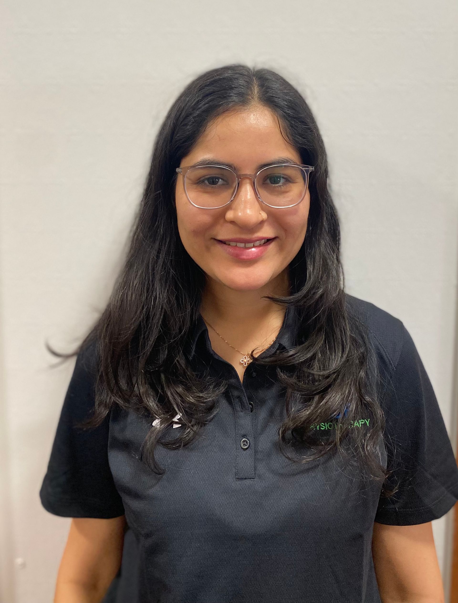 Woman with glasses smiles, wearing black shirt against a white wall.