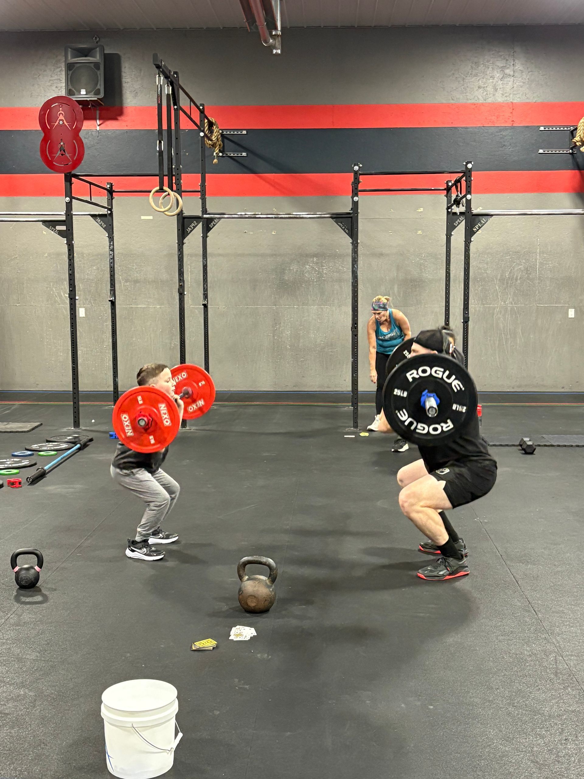 A coach instructs a group of people in a gym as they hold wooden dowels and practice squat form.