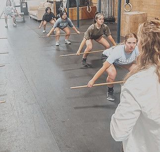 A coach instructs a group of people in a gym as they hold wooden dowels and practice squat form.