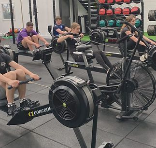 A coach instructs a group of people in a gym as they hold wooden dowels and practice squat form.