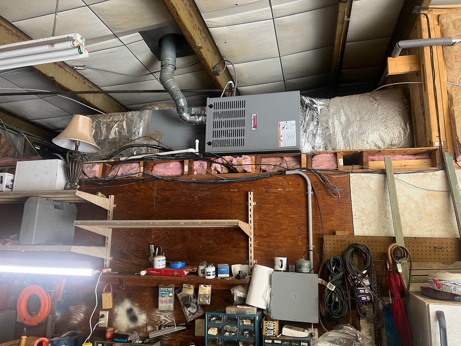 Garage interior with HVAC unit, ductwork, tools, shelves, and exposed insulation.