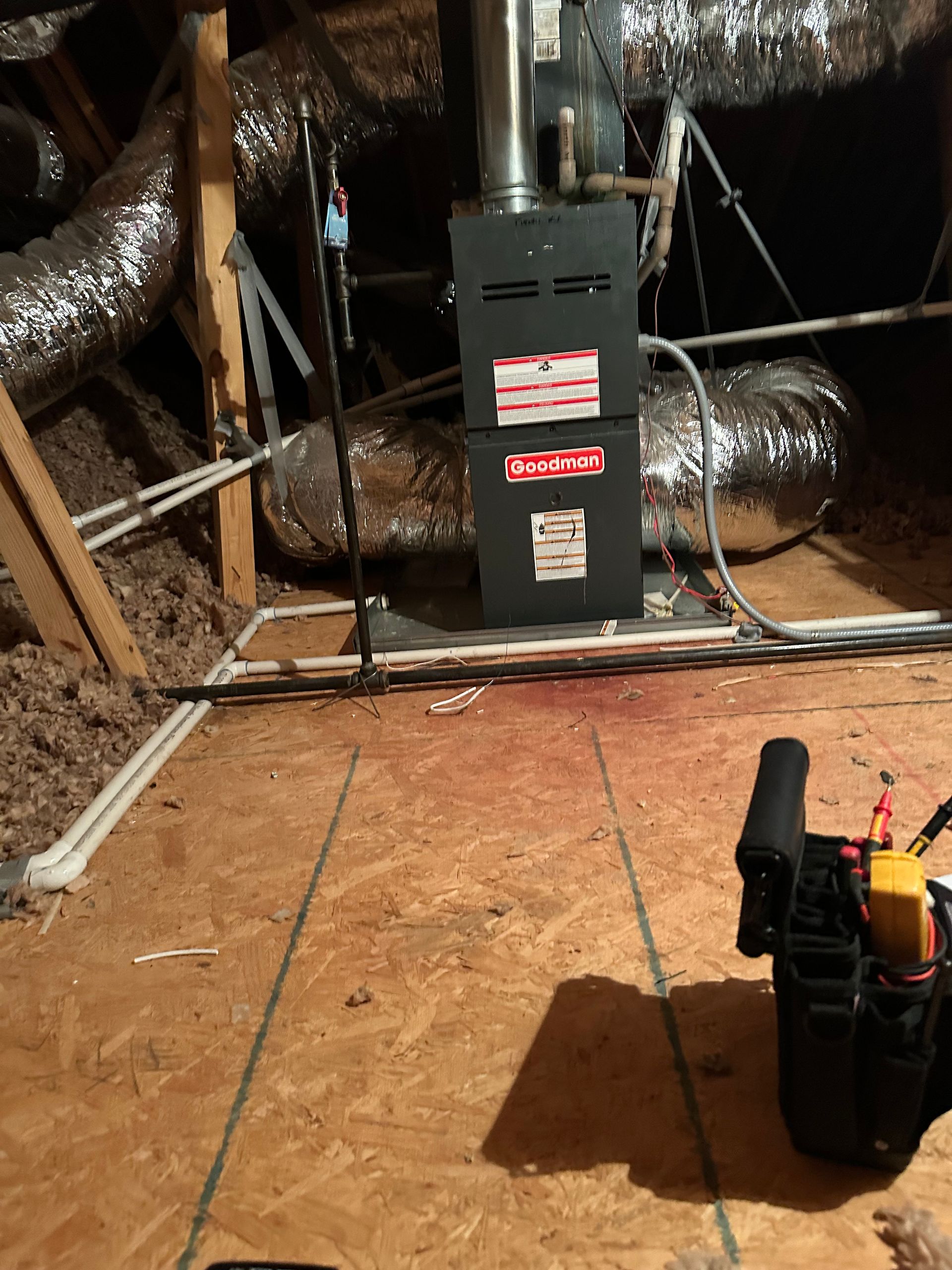 Furnace in an attic. Ductwork, wood flooring, and tool bag present.