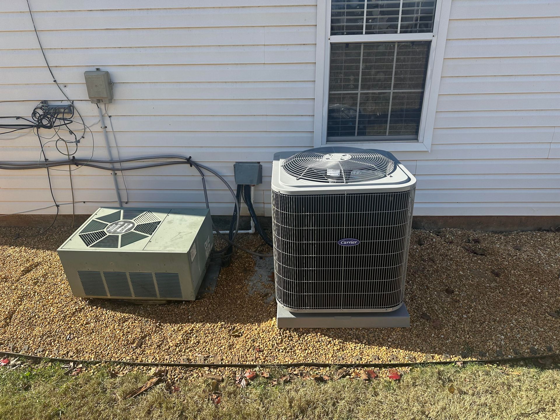 Two air conditioning units sit outside a building with a window, on gravel.