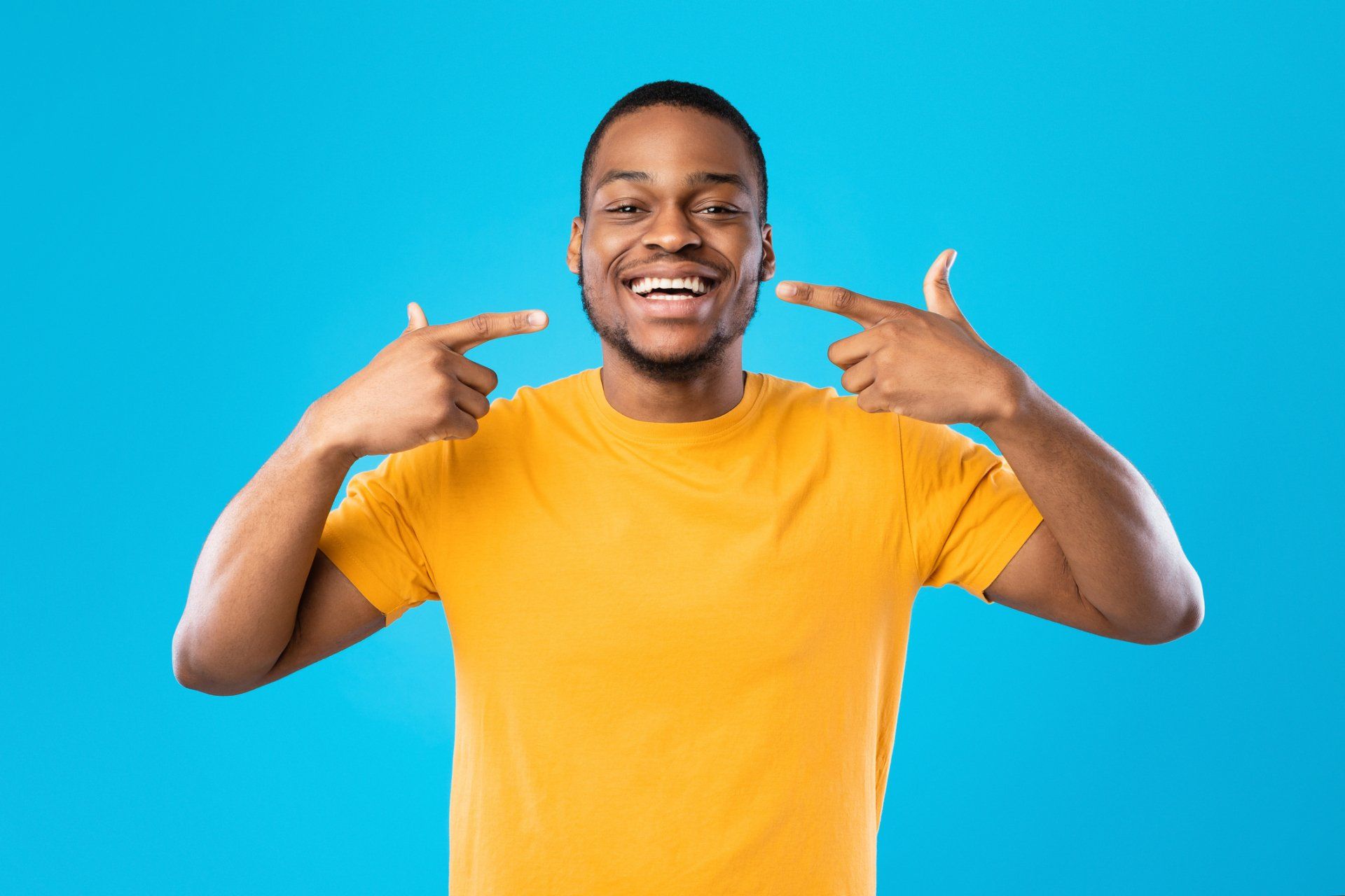 Man with a bright smile pointing at his teeth, wearing a yellow shirt, set against a blue backdrop.