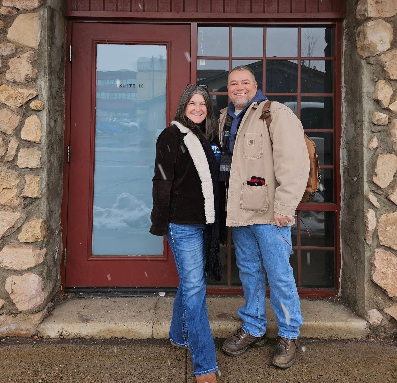 Couple stands outside a building with stone walls. The woman wears a brown jacket, jeans. The man wears a tan coat, jeans. Snow is falling.