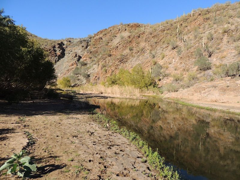 A calm body of water reflects the surrounding rocky hills and sparse vegetation under a clear blue sky.