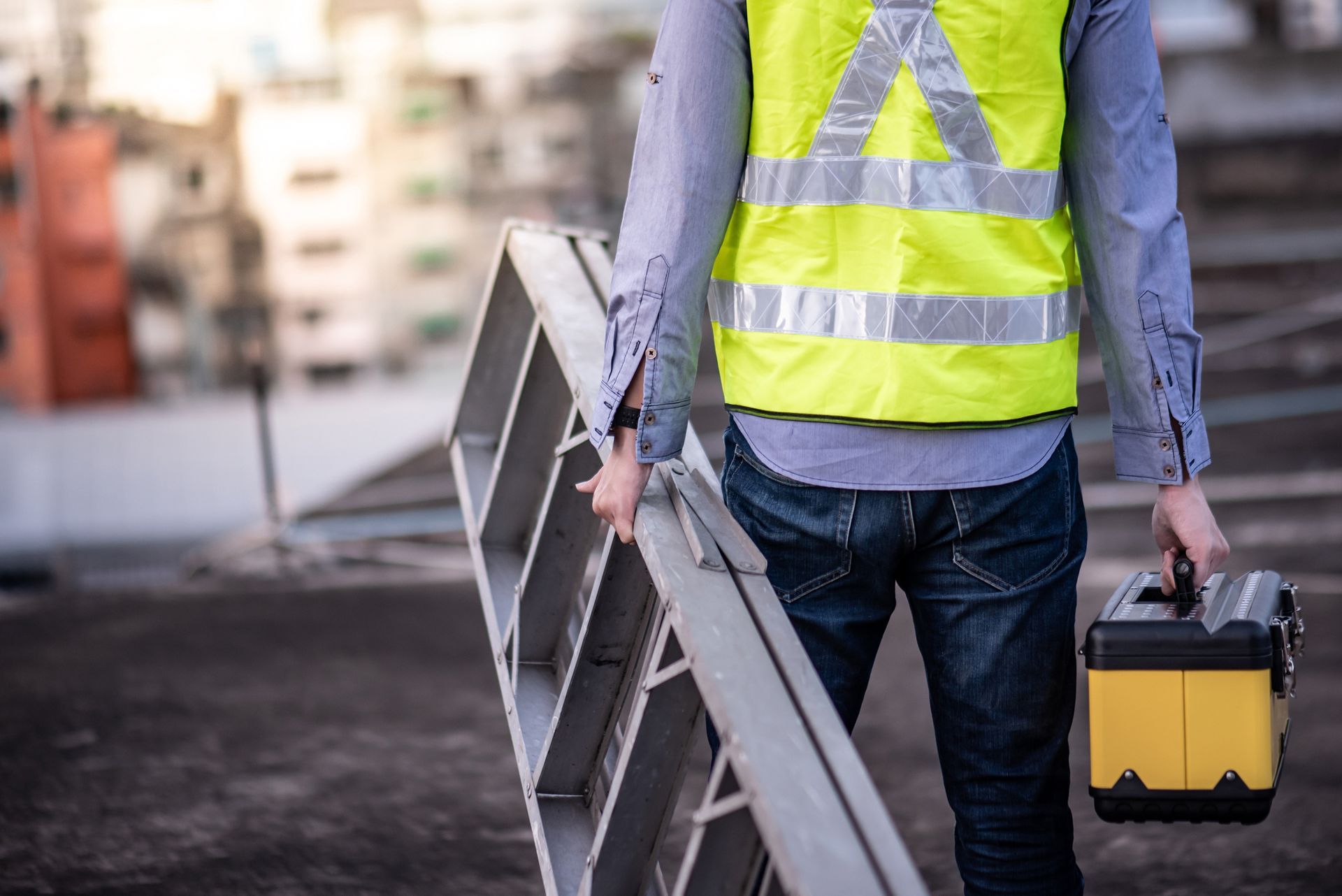 Person in a yellow safety vest carrying a toolbox and ladder on a rooftop.