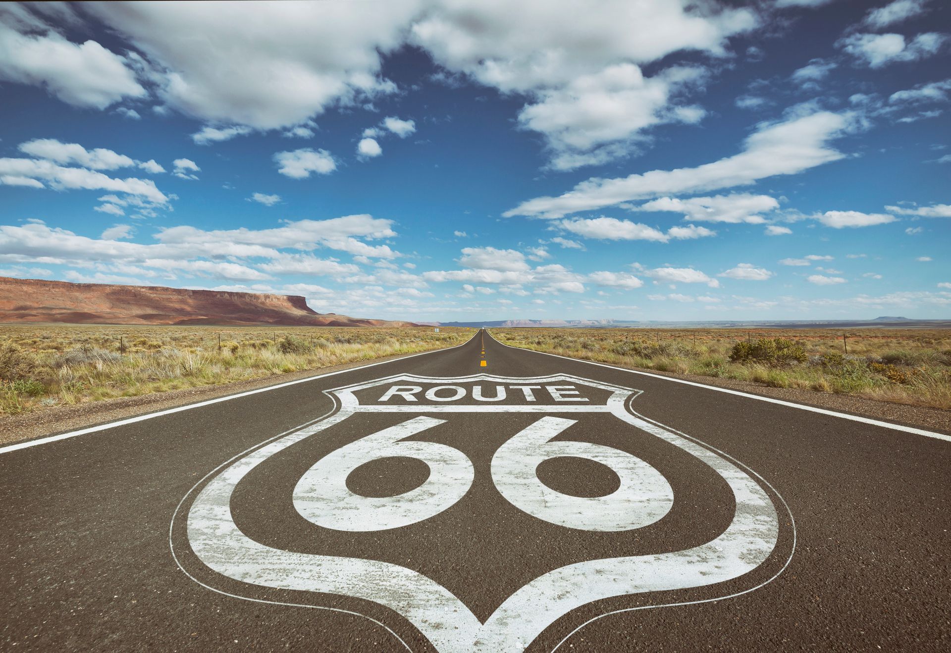 Route 66 sign on asphalt road, leading toward a horizon, with blue sky and clouds overhead.