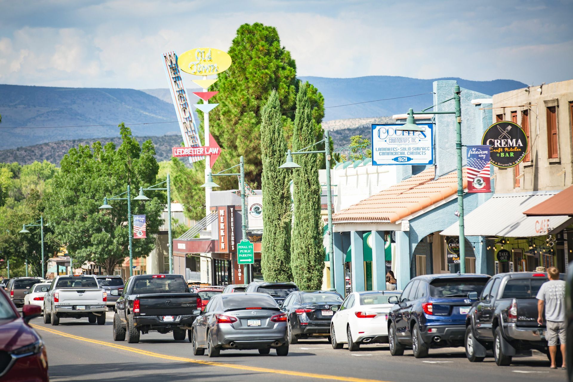 Street view with cars and shops, green trees and mountains in the background.